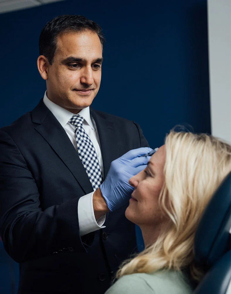 Dr. Bhrany, wearing a dark suit and blue nitrile gloves, administers an injection to a woman's forehead. The woman, with wavy blonde hair and eyes closed, is seated in a medical chair with a dark blue headrest. A syringe is visible in Dr. Bhrany’s gloved hand. The setting suggests a medical aesthetic procedure, such as Botox. The background is light and neutral, with part of a wall visible. - Botox in Seattle, WA