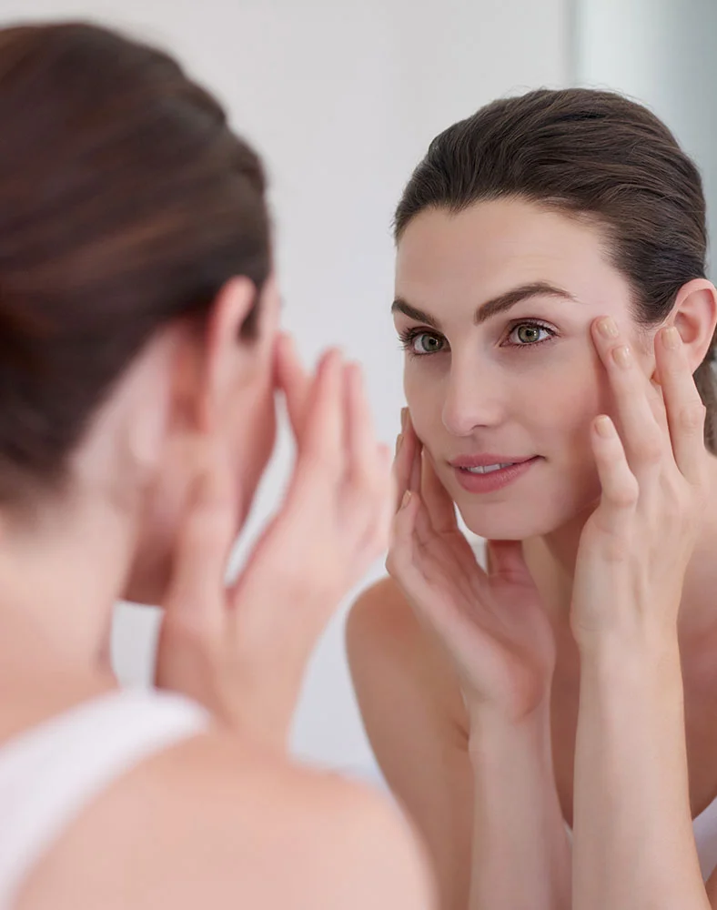 A woman with shoulder-length brown hair looks at her reflection in a mirror, touching her face with both hands as she examines her skin. She wears a simple white tank top and has a focused expression. The background is a light, neutral space, likely a bathroom or vanity area, with her reflection clearly visible in the mirror. - Brow Lift in Seattle, WA