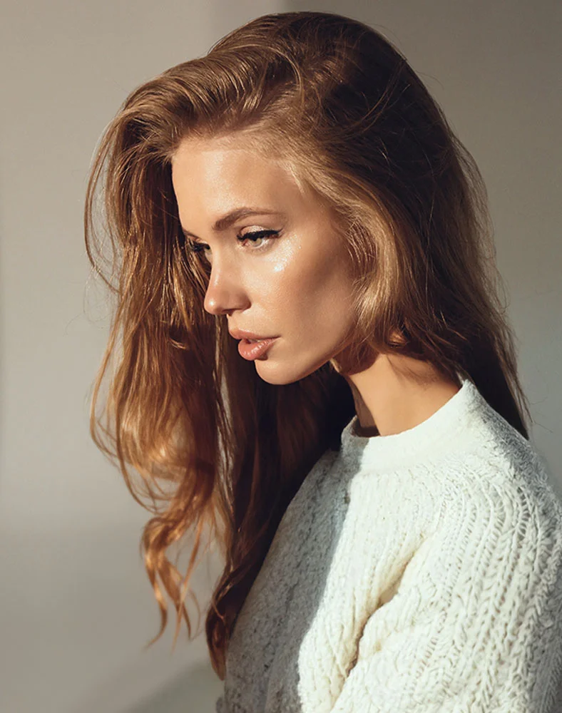 Close-up of a woman with long, light brown, naturally wavy hair falling loosely around her shoulders. She has light skin and a thoughtful expression, with her head slightly turned to the right and gaze directed downward. Her face features a delicate nose, full lips, and a subtle glow. She is wearing a cream-colored knitted sweater with a visible textured pattern. The background is a simple, light beige or gray tone, and soft natural lighting casts gentle shadows on her neck and shoulders. - Buccal Fat Removal in Seattle, WA