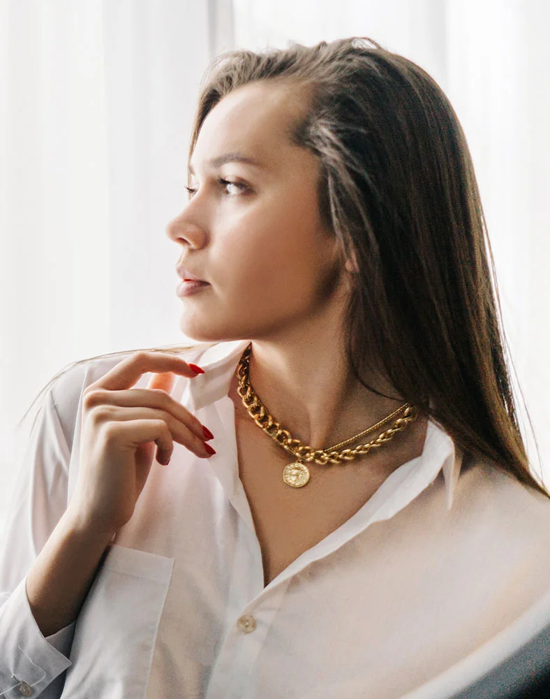 Profile view of a young, light-skinned woman with shoulder-length dark brown hair. She wears a white button-down shirt and a thick gold chain necklace with a medallion pendant. Her hand, with red fingernails, touches her shirt collar. The background is a soft white sheer fabric, with diffused light gently illuminating her face and body. - Chin Augmentation in Seattle, WA