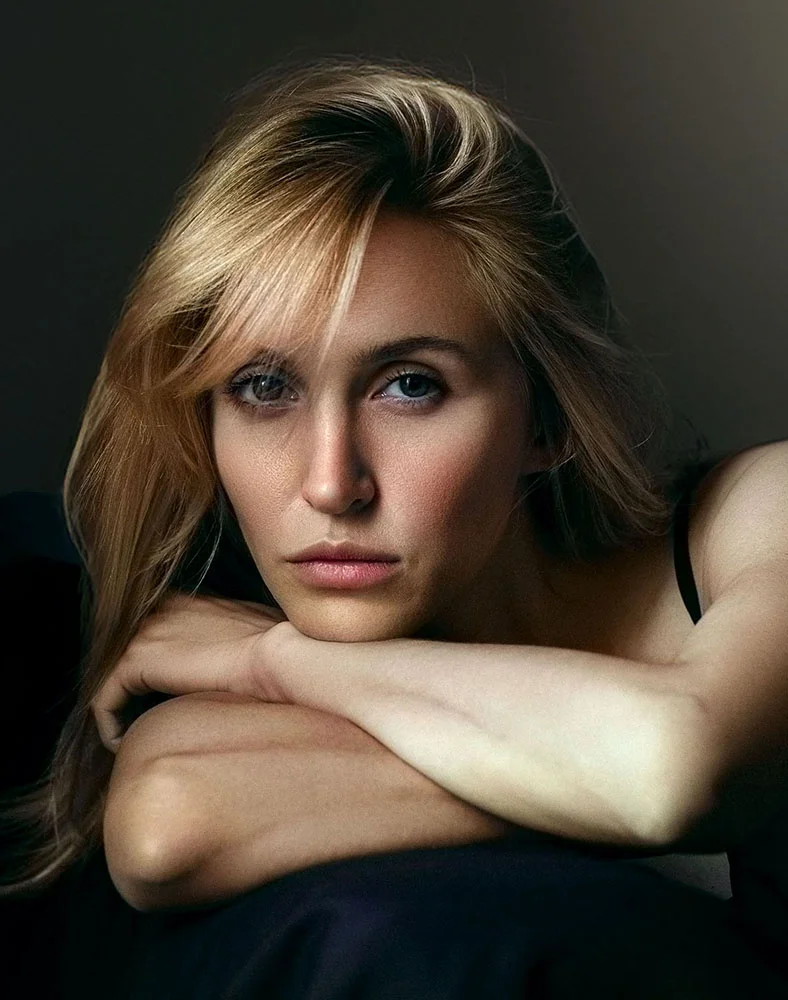 A light-skinned woman with shoulder-length blonde hair is seated, resting her head on her arms. She wears a dark sleeveless top and gazes slightly downward with a serious, thoughtful expression. The image has a low-key, moody lighting and color palette, with a muted dark gray-brown background and her arms resting on a dark surface. - Diamond Glow in Seattle, WA