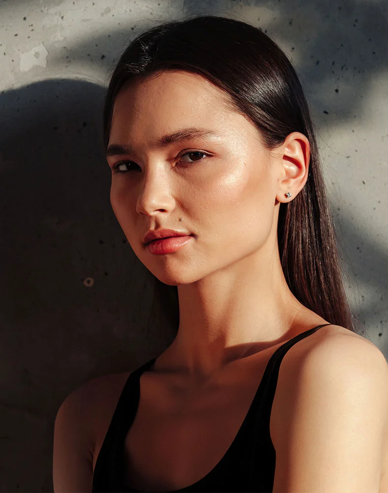 A woman with long dark hair stands against a textured wall, illuminated by soft sunlight with shadows cast on the surface behind her. - Earlobe Repair in Seattle, WA