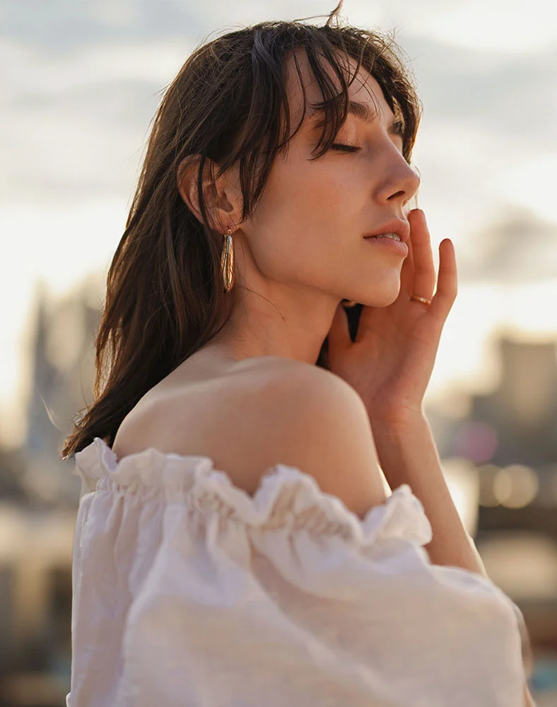 A close up of a woman with dark brown hair from the shoulders and up. She is wearing big gold earrings as she glances down away from camera. - Eyelid Surgery in Seattle, WA