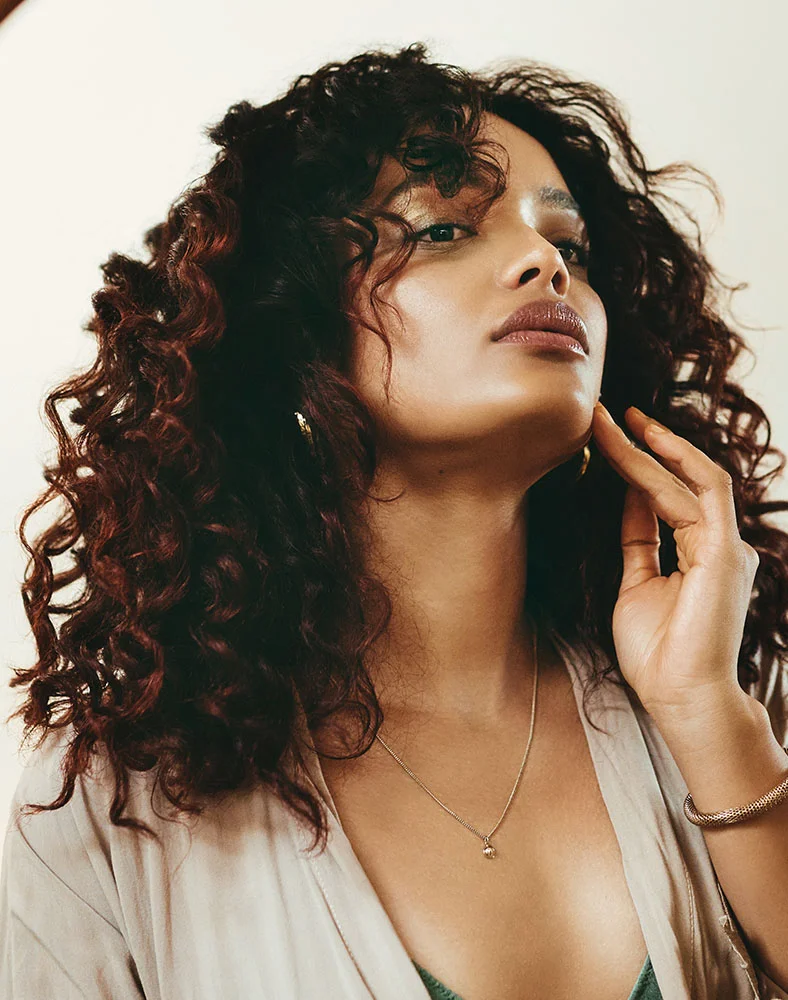 Close-up portrait of a woman with dark, curly hair and light brown skin, looking slightly upward and to her right with a calm, thoughtful expression and a gentle smile. She wears muted mauve-brown lipstick and light golden eyeshadow. A delicate light-gold necklace with a small pendant rests on her neck. Her hand gently touches her neck and jawline. She is dressed in a loose, light beige or cream-colored top with a hint of a teal-green top underneath. The soft, light beige background enhances the warm, natural mood of the image. - Laser for Skin Pigmentation in Seattle, WA