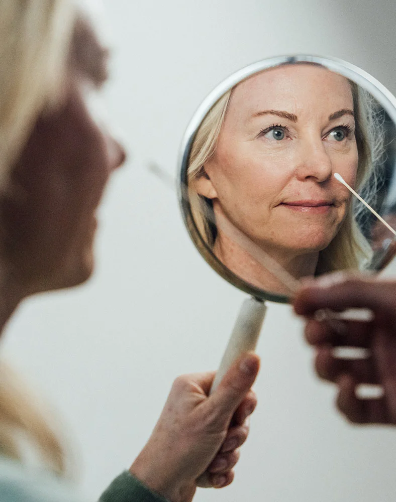 A close up shot of a blonde woman holding a mirror as the doctor uses a swab to point to her face. The woman is seen clearly in the mirror with her side of her face blurred. - Nasal Surgery for Breathing in Seattle, WA