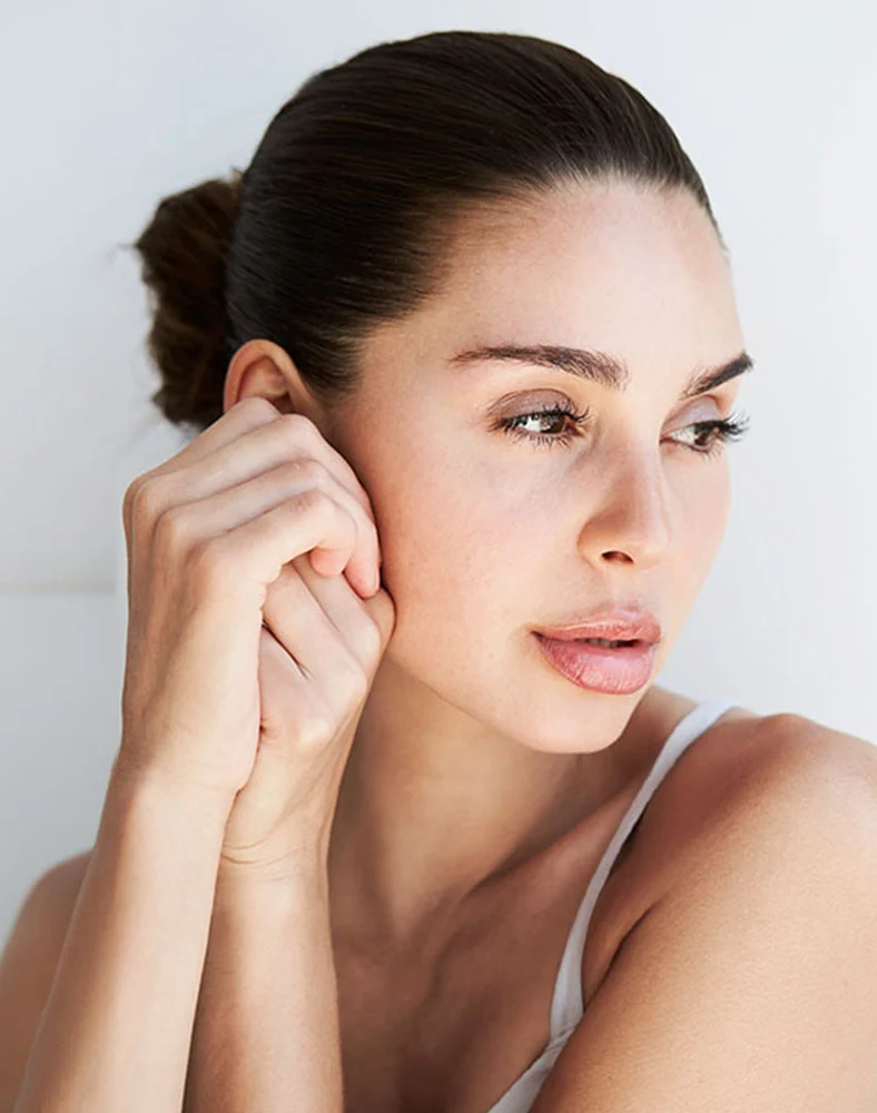 Close-up of a woman with dark brown hair pulled back in a bun, revealing her smooth, light-toned skin. She gazes slightly off-center with a calm, attentive expression. Her well-defined eyebrows, light pink lips, and softly parted smile highlight her natural features. The plain white background keeps the focus on her face and upper chest. - RF Microneedling in Seattle, WA