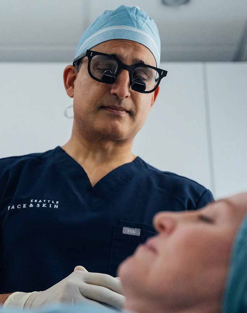Dr. Bhrany, wearing a navy blue scrub top with "SEATTLE FACE & SKIN" embroidered on the chest, looks down at a woman lying on a table. He wears a light-blue surgical cap, white medical gloves, and black magnifying glasses. The woman, with eyes closed and also in a light-blue surgical cap, lies on her back in a light-colored medical office or clinic. - Understanding Aging Faces in Seattle, WA